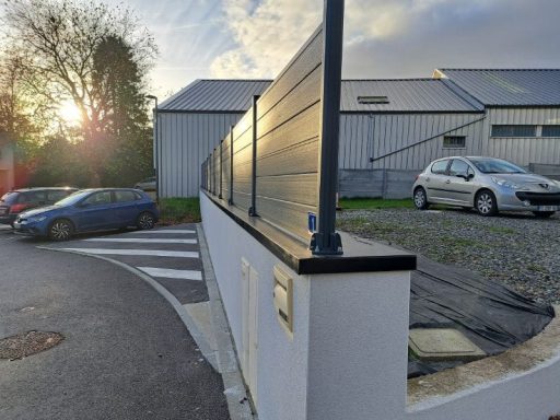 Muret en béton avec brise vue, voitures garées et soleil couchant à l'arrière.
